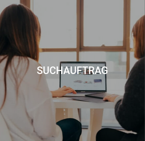 Zwei Personen sitzen an einem Tisch und arbeiten gemeinsam an einem Laptop, der Text "SUCHAUFRAG" ist über dem Bild positioniert.